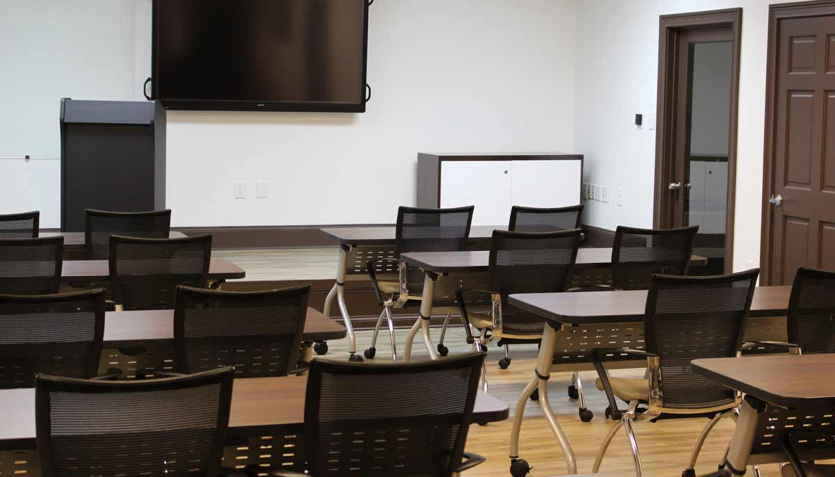 Atlanta Exec Suite's Training facility, with wooden tables that can be arranged and executive style training chairs.  The photo is from the back aisle up toward the raised stage with a podium and large screen television.